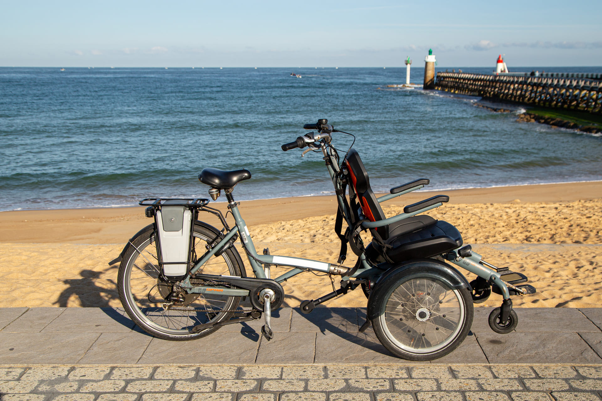 Photographie d’un vélo adapté avec siège et roue arrière élargie stationné face à la mer, sur une promenade en bord de plage avec digue et phare à l’horizon